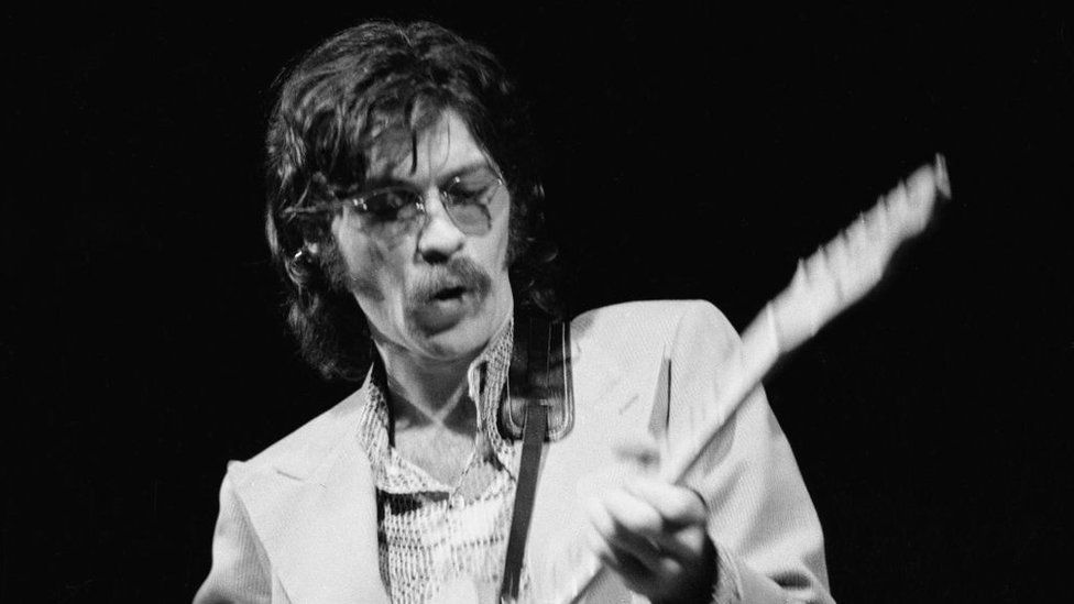 Canadian instrumentalist  Robbie Robertson performing with The Band astatine  the Royal Albert Hall, London, 3rd June 1971. He's wearing a achromatic  overgarment   and holding an electrical  guitar.