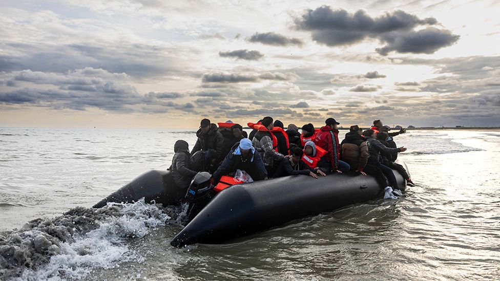 Migrants in lifejackets sit on board a small smuggler's boat with a dramatic cloudy dawn skyline in the background, in an attempt to cross the English Channel, on the beach of Gravelines, near Dunkirk, northern France on April 26, 2024