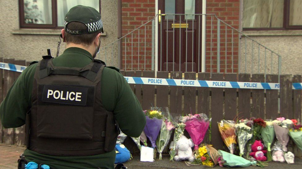 A police officer stands next to a police cordon outside a house where floral tributes to Ali Maguire are laid
