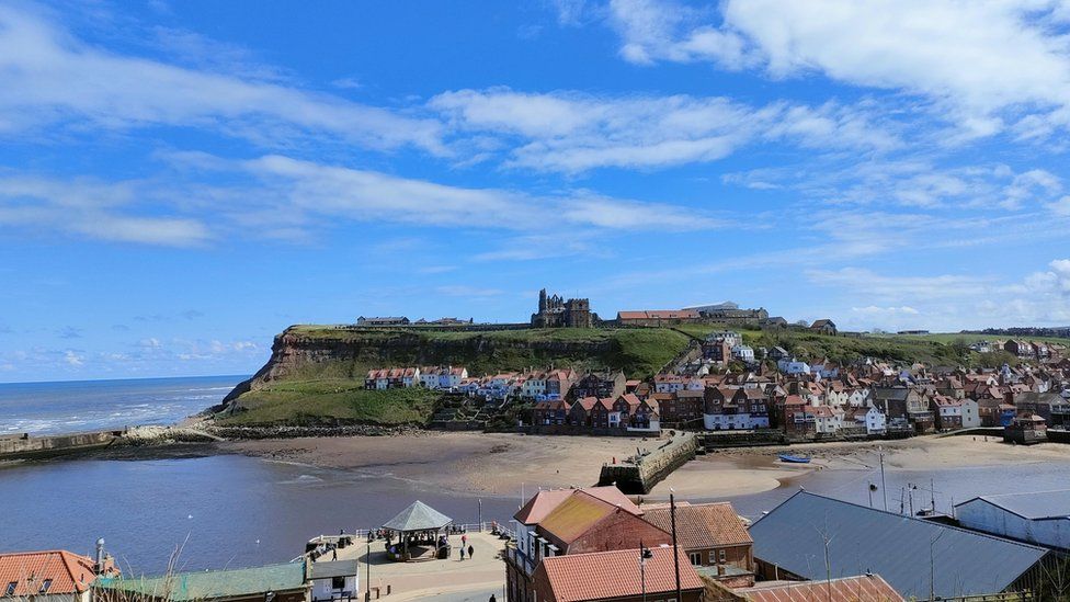 Photograph of a beach and Whitby castle on top of the hill across the bay. Blue skies and a few white clouds.