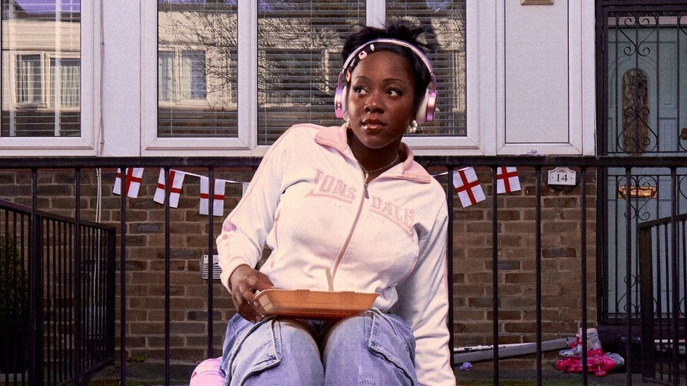 Rachel Chinouriri. Rachel is a 25-year-old black woman with long dark hair worn tied back with her side fringe secured with five multi-coloured clips. She wears a pink and white Lonsdale tracksuit top and sits outside a London flat decorated with St George's cross bunting. She has a polystyrene takeaway container on her lap and wears enormous pink headphones on her head.