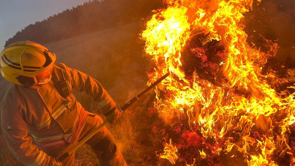 A firefighter in full protective equipment prods a large fire in a field