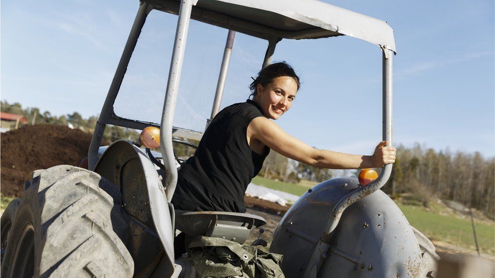 A brunette woman sat on a tractor looking over her shoulder at the camera, with one hand resting n the side bar.