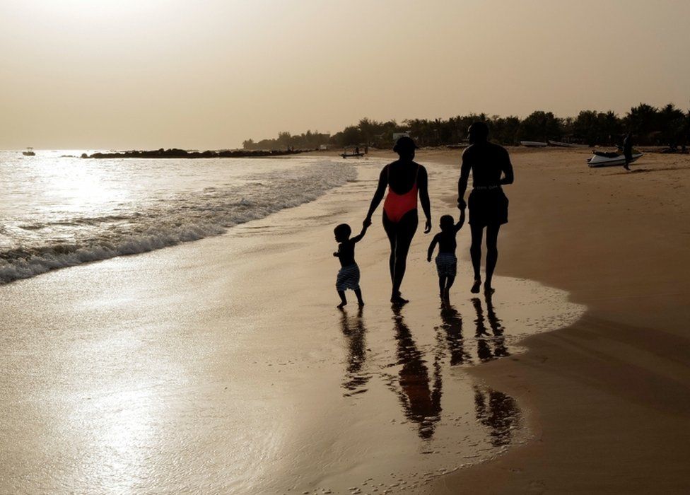 A man, woman and twin boys dressed in bathing suits walk along a beach.