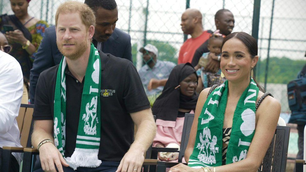 Harry and Meghan watch a game of sit-down volleyball wearing Nigerian scarves