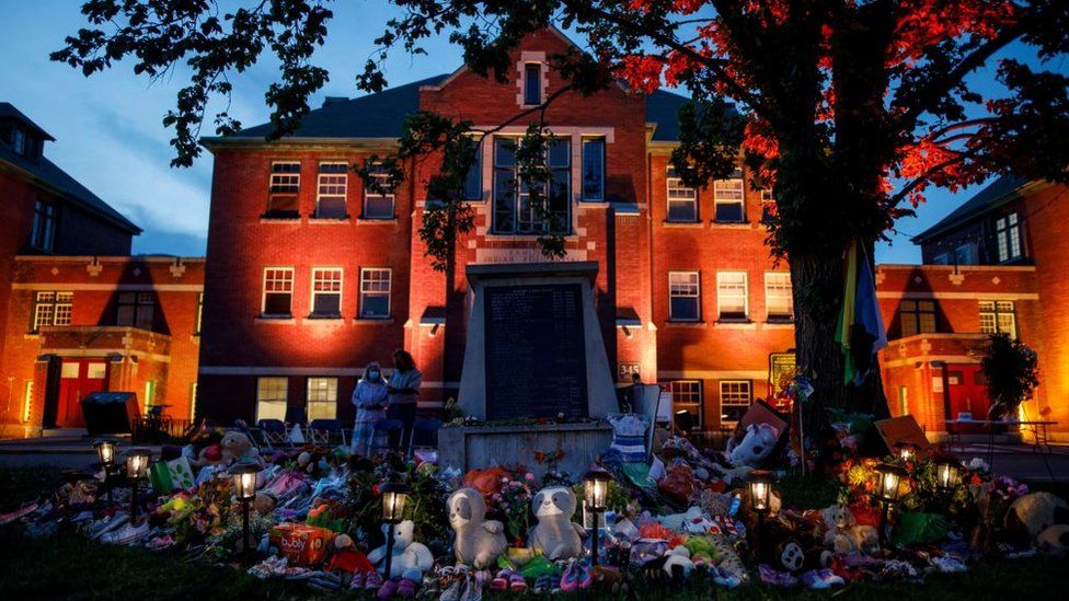 Импровизированный мемориал в честь 215 детей, чьи останки были обнаружены в захоронении рядом с объектом, виден в оранжевом свете, освещающем фасад бывшей школы-интерната для индейцев Камлупс в Камлупсе, Британская Колумбия, Канада, 2 июня 2021 года.