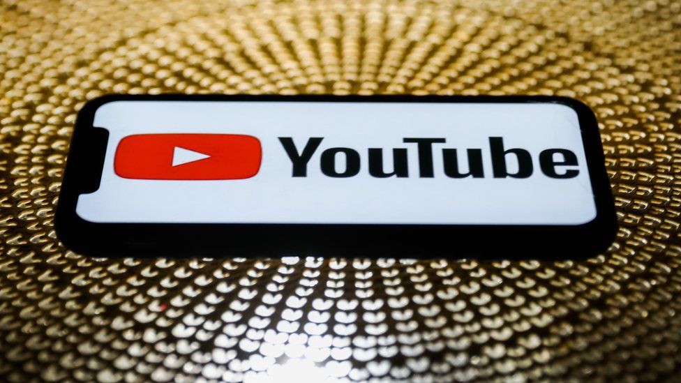 YouTube logo is seen on a smartphone laid down on a table, radiating a gold substance - which could be sequins, or coins - in a circular pattern around it