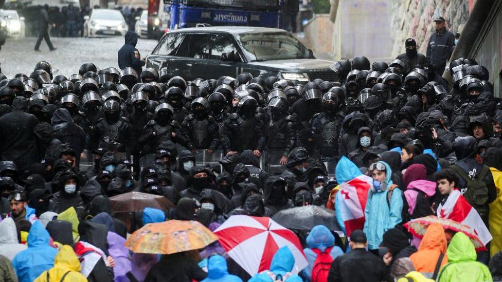 A mass of police in black block a road, and protesters in colourful raincoats gather nearby