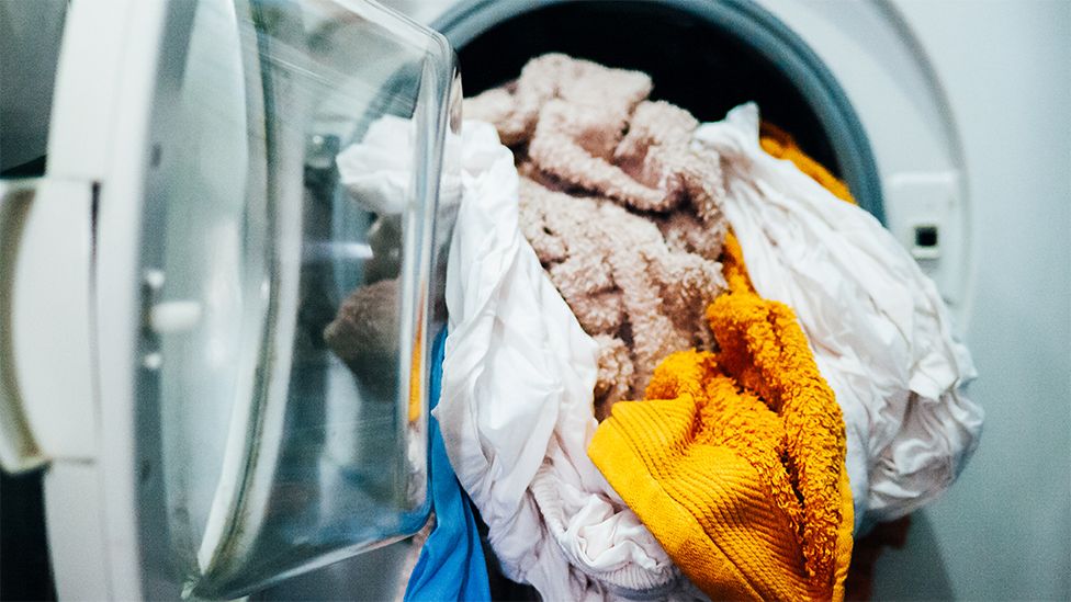 A white washing machine with the door open, and a pile of towels hanging out. The towels are orange, brown and there are sheets there as well.