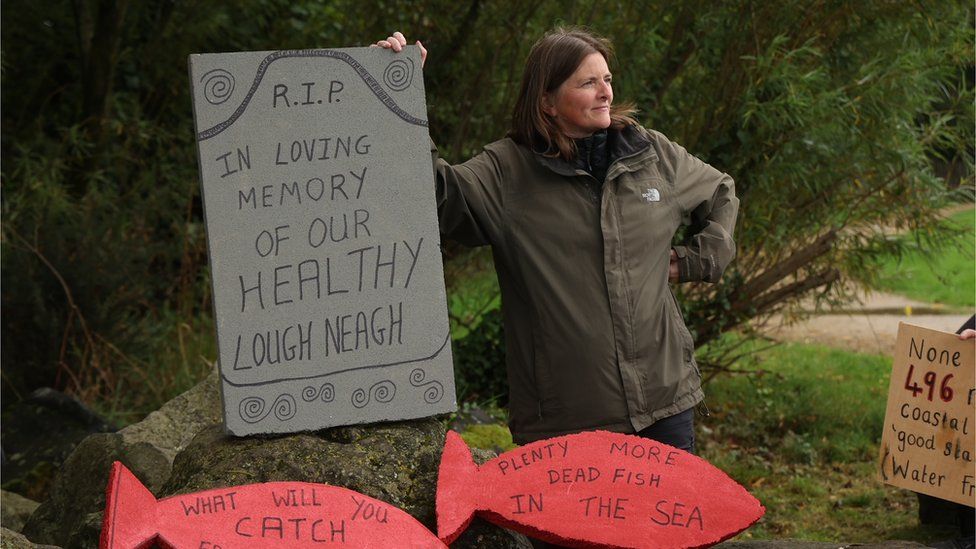 Woman stood with headstone reading 'In memory of our healthy Lough Neagh'