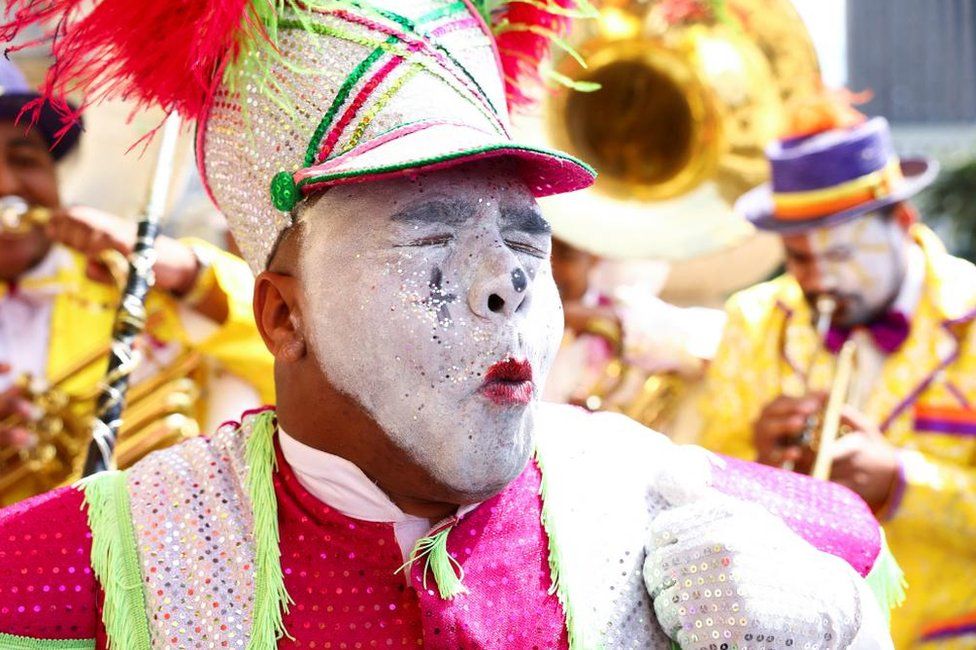A man wearing white face paint with a colourful elabourate hat and pink outfit. His eyes are closed and he is making an expressive expression.