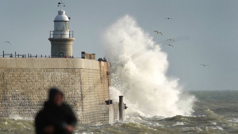 A fisherman on the beach in Folkestone, Kent, as waves crash against the harbour wall during strong winds