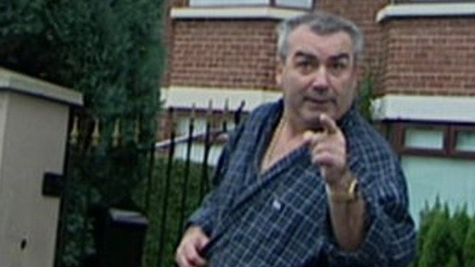 Freddie Scappaticci pointing at the camera with his eyebrows raised. Semi-detached houses and a hedge are visible behind him.