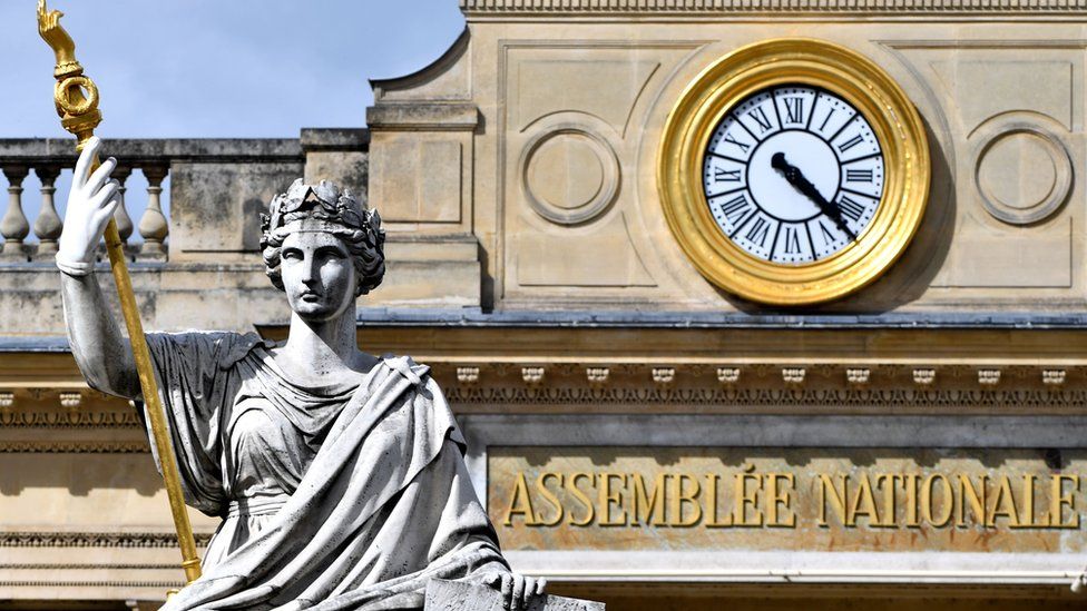 The French National Assembly is seen in this close-up photograph, framing the sign and golden clock on the facade and also the statue of a woman holding a golden sceptre which stands before the entrace