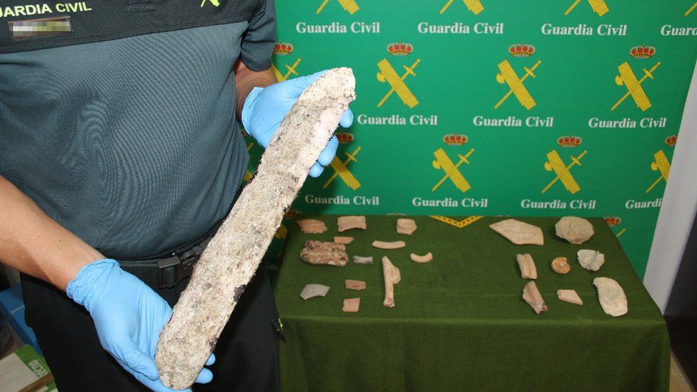 A gloved officer holds a long, thin piece of rough stone in front of a table littered with smaller fragments