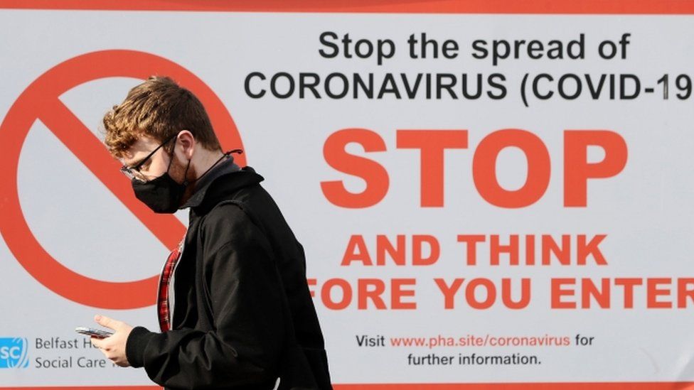 A young man wearing a mask looking at his phone and walking in front of a Covid-19 awareness poster from the Department of Health