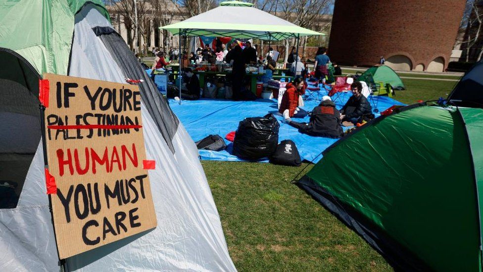 Part of an encampment at the Massachusetts Institute of Technology. A placard on the side of a tent in the foreground reads: "If you're Palestinian, you must care". The world 'Palestinian' is then crossed out and replaced with the word 'human'.