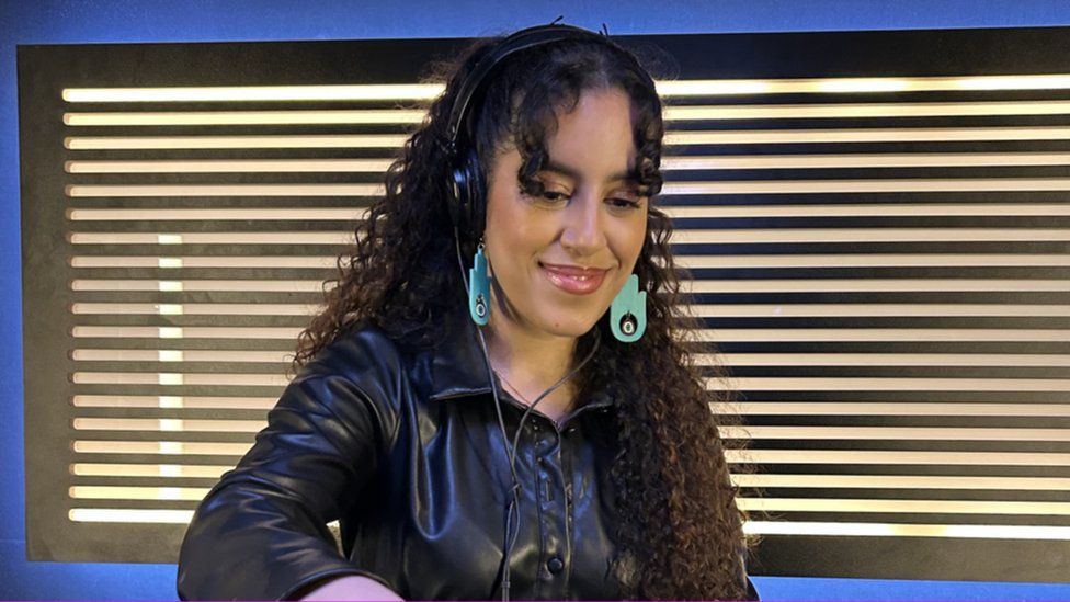 Nooriyah smiles as she mixes in a recording studio. She's got shoulder-length brown curly hair, with large, dangly cyan-coloured earrings in a Hand of Fatima design. That's a flat with an eye in the middle of the palm. Nooriyah's also wearing over-ear headphones and a black leather jacket.
