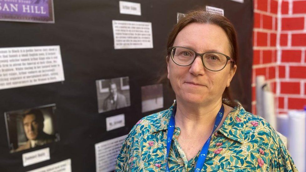 Woman with short brown hair tied back, and glasses stands next to a library display board