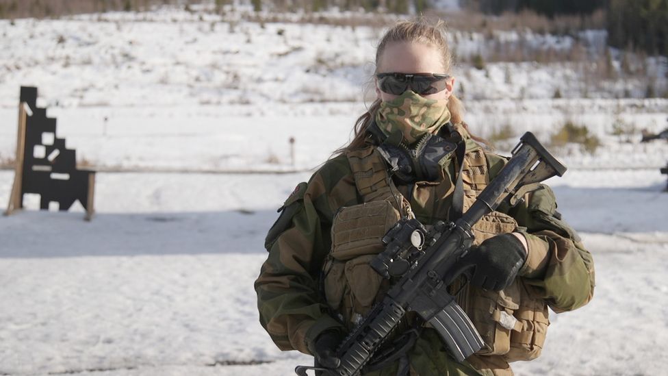 Jannike holding a gun and looking at the camera. She wears sunglasses and a mask covers her nose and mouth