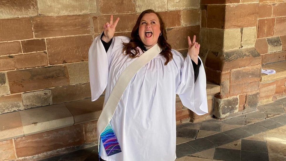 Rev Jessica Fellows, a woman wearing a white robe, with a "peace sign" of two fingers raised on each hand. The wall background shows brown bricks.