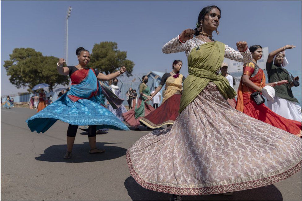 ผู้หญิงสวมชุดอินเดียพื้นเมืองสีสันสดใสเต้นรำ  พวกเขาอยู่ข้างนอกและมีท้องฟ้าสีครามและต้นไม้อยู่ข้างหลังพวกเขา