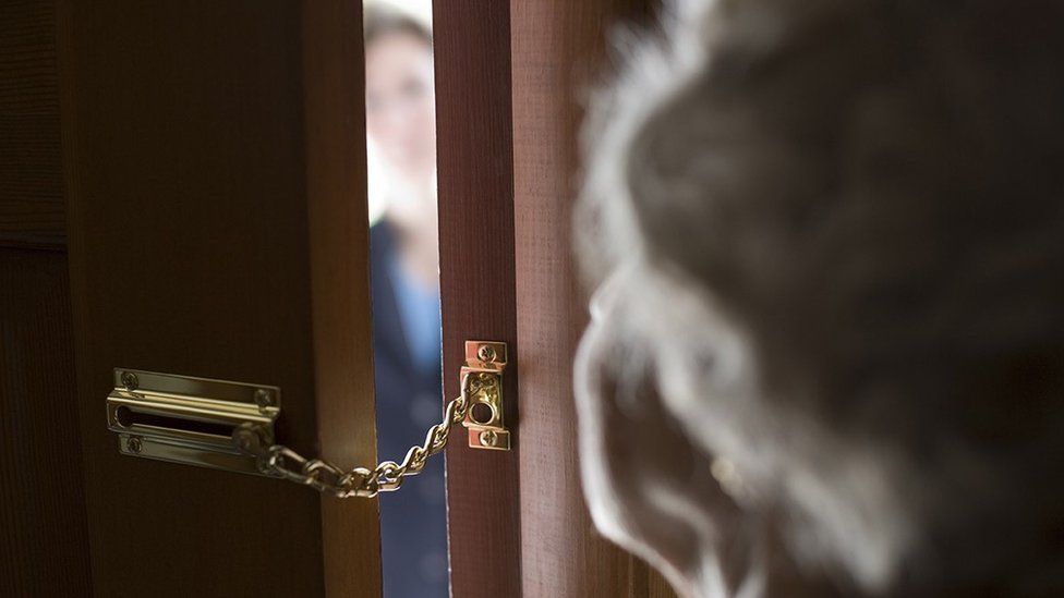 An elderly person answering a door with a chain on it