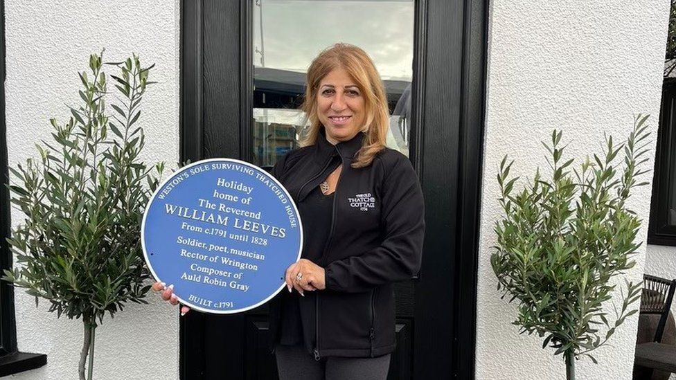 Maria Michael holding a blue plaque outside the Old Thatched Cottage