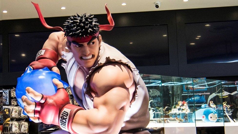 A life-size statue of Ryu from Street Fighter, in his iconic white karate suit and red bandana, prepares a "hadouken" energy ball. In the background is a Capcom brand store