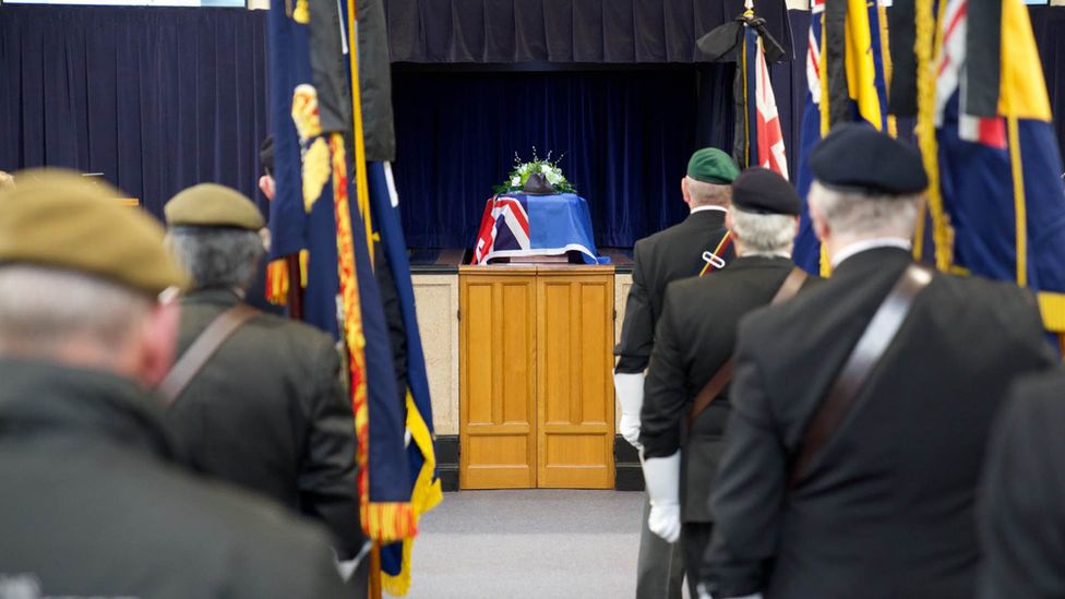 The aisle of the crematorium with the coffin draped in a flag. On either side of the crematorium veterans and serving soldiers stand.