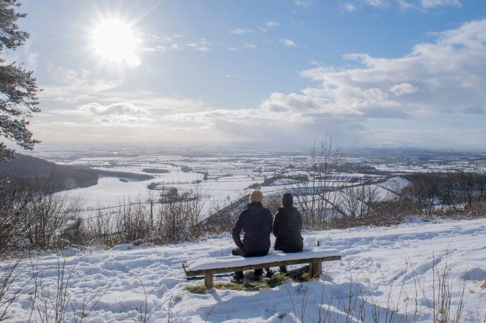 A couple sit on a bench overlooking the snow-covered fields of Thirsk at Sutton bank National Park Centre in the North Yorks Moors National Park in North Yorkshire