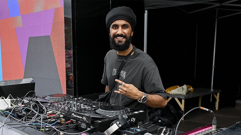 Virdi, an Asian man wearing a black turban representing his Sikh faith. wearing a grey tshirt. He's on DJ decks with multiple wires, buttons and faders.