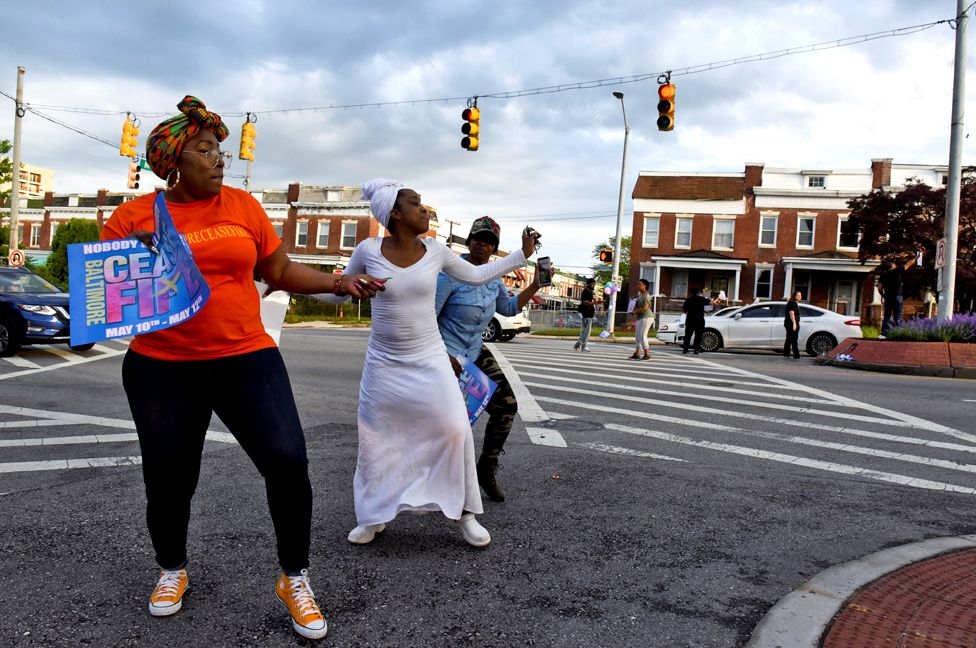 Люди танцуют на улице с плакатами с надписью «прекращение огня» в Балтиморе. Ceasefire — это местная организация, которая проводит мероприятия, чтобы привлечь внимание к вооруженному насилию в Балтиморе