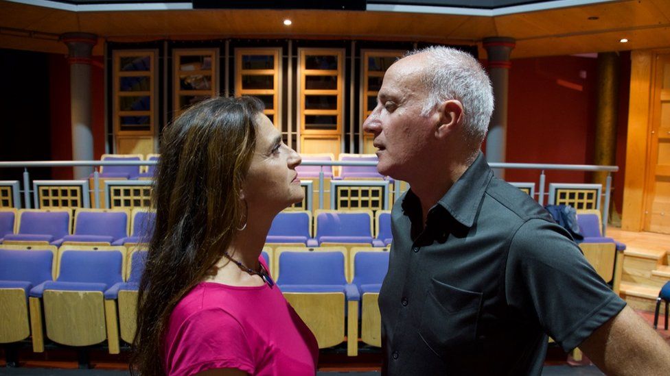 A woman and a man stare each other down, nose to nose, in a theatre rehearsal room