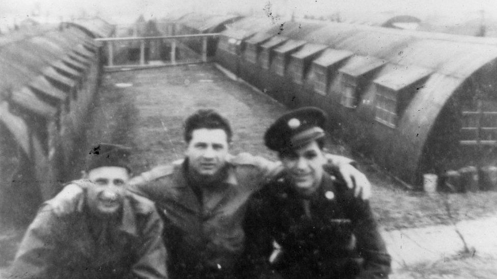 Black and white photo of three soldiers in front of nissen huts
