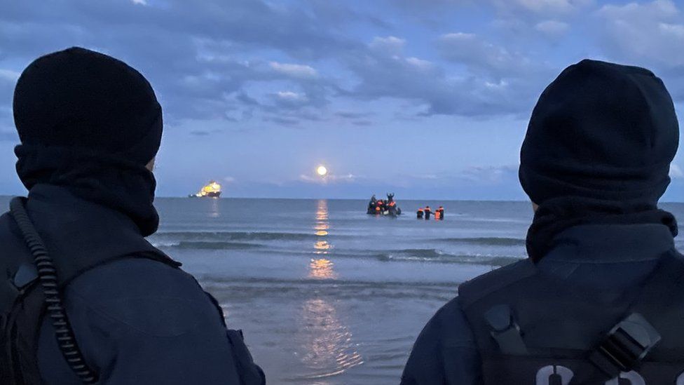 Officers look out to sea as migrants board boat