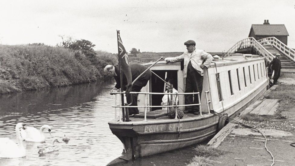 Black and white photograph showing Lord Rusholme, another man and a woman at the stern of the boat, Lord Rusholme is feeding some swans. The boat is moored on the canal, there are also people visible at the bow. There is a footbridge and a building in the background.