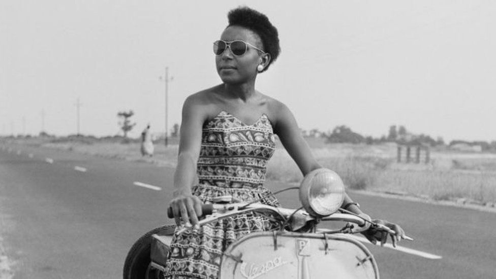 A woman sitting on a moped by the roadside poses for the camera.