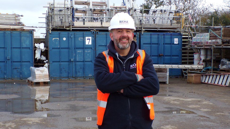 Image of Paul Barnsley. He standing on a construction side with scaffolding behind him. He is wearing black trousers, black quarter zip and a orange high-vis jacket with a white helmet. He is smiling at the camera with his arms crossed.