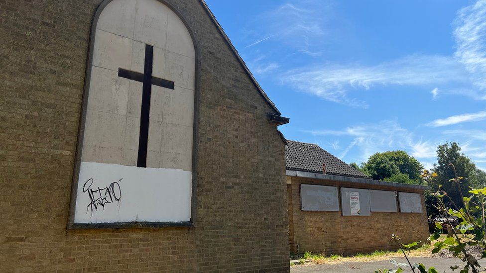 A modern brick-built church, with windows boarded up and graffiti showing