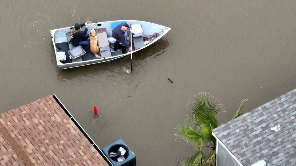 A man rows a boat with a woman and a dog in floodwater
