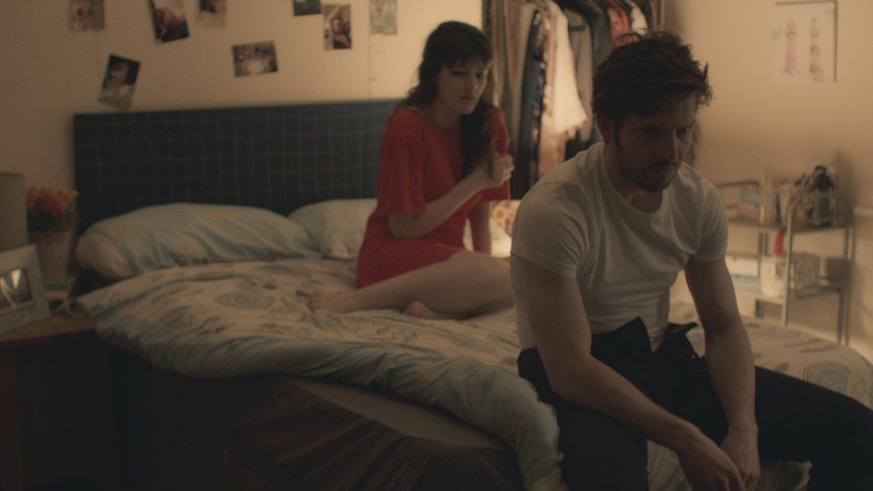 A still from a film. A man sits on the edge of a bed, behind him a woman lies on the bed.
