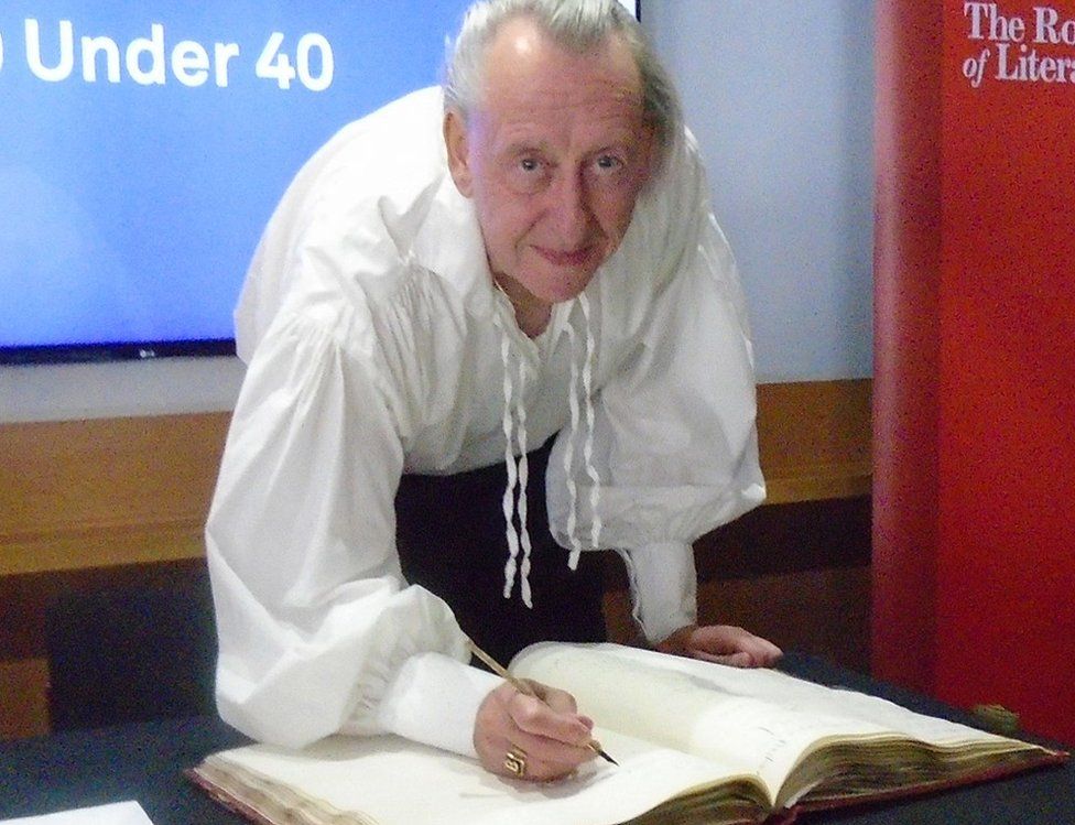 A man in a large white shirt signs a large book with an old pen