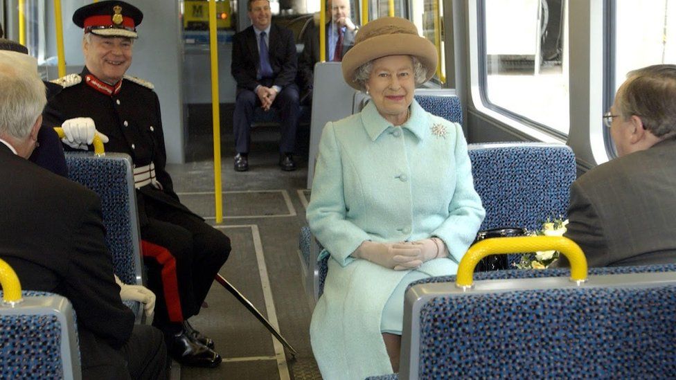 The Queen sits on a seat on a Metro Carriage
