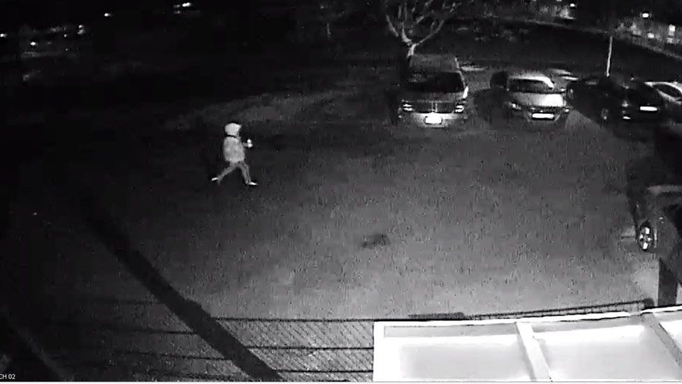 CCTV shot of a person wearing a cap or hood walking past parked cars