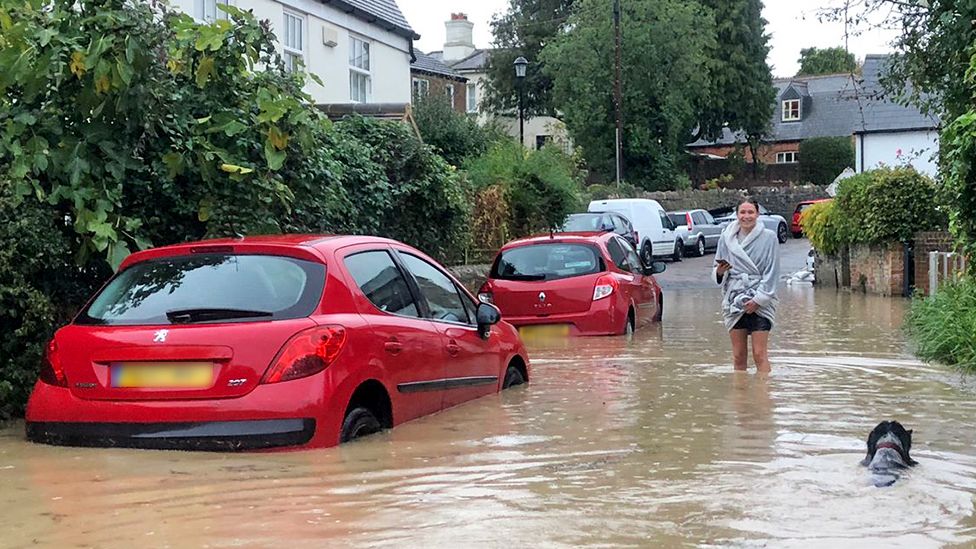 Flooding in Newport, showing a flooded road with two red cars and a person on their phone in a rain mac and a black dog wading through the water