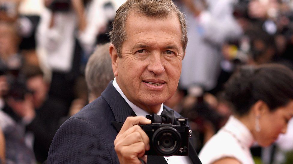 mario testino holds a camera on a red carpet