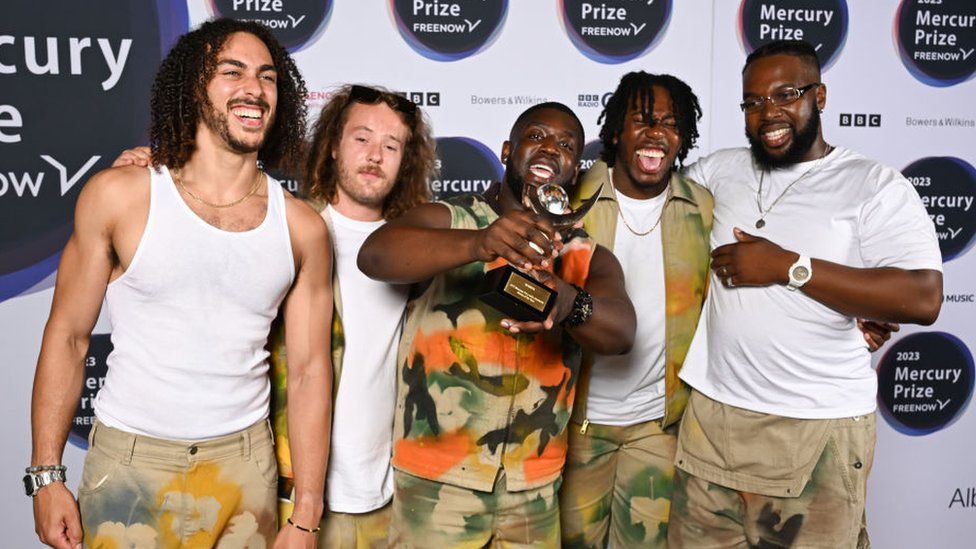 Members of Ezra Collective, (L-R) James Mollison, Joe Armon-Jones, Femi Koleoso, Ife Ogunjobi and TJ Koleoso after winning The Mercury Prize during The Mercury Prize 2023 awards show at Eventim Apollo on September 07, 2023 in London. The group all wear white T-shirts and camouflage-style trousers except for Femi, who wears the same pattern on his top. Femi is holding the trophy towards the camera and all members of the group are smiling.