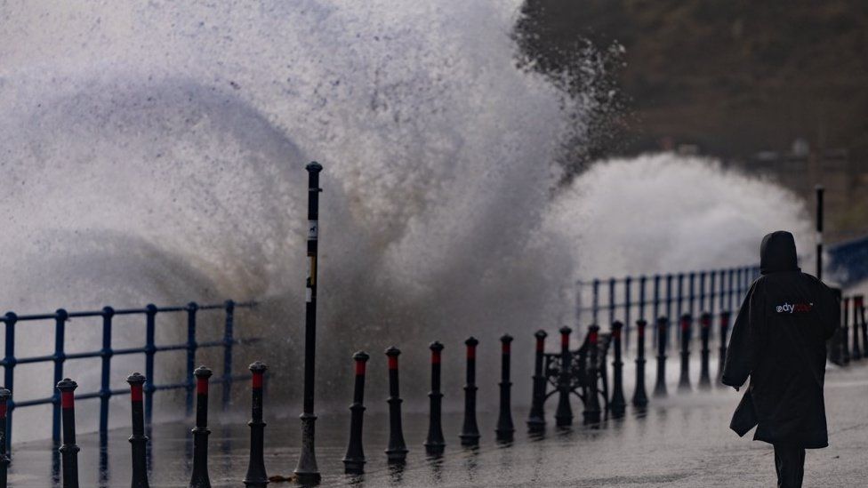 A woman stands on a seaside promenade as waves crash over
