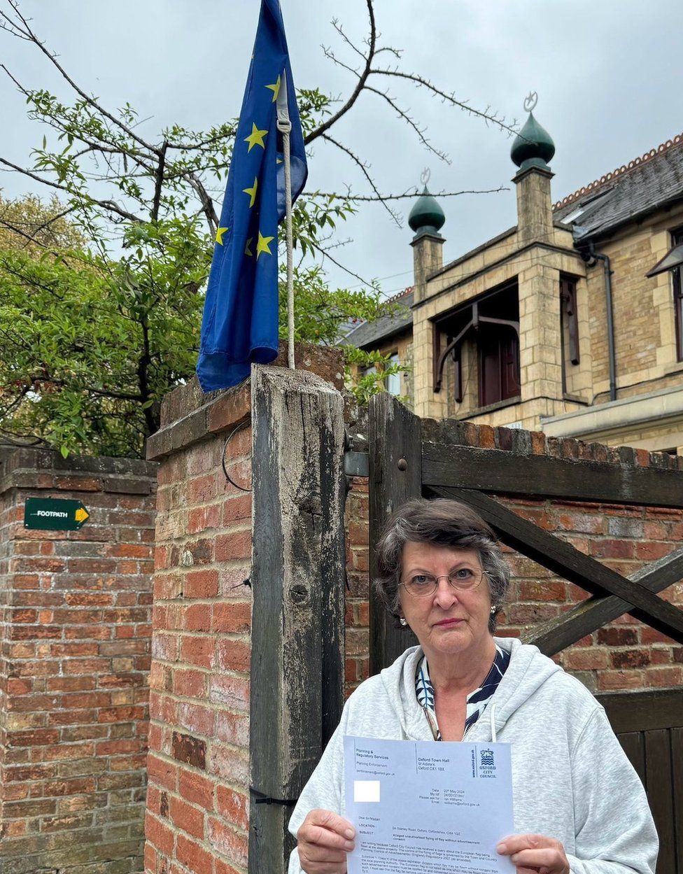 Catherine Bearder, who has short greying, brown hair and is wearing a pale grey, hooded sweater, thin-rimmed round glasses and pearl earrings, pictured outside her home with a Council of Europe flag flying from the top of a tall wooden gatepost. There is also a high brick wall with a tree on the other side